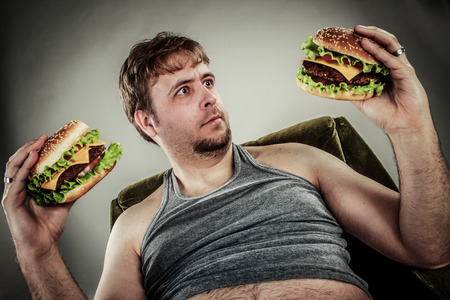 Fat man eating hamburger seated on armchair. Style fast food.の写真素材