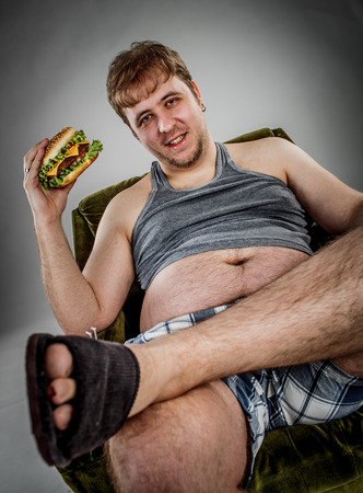 Fat man eating hamburger seated on armchair. Style fast food.の写真素材