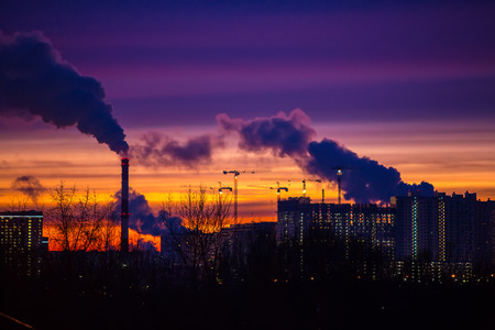 Modern city in the evening at sunset. Smoke comes out of the pipes of power plants and factories of the modern neighborhood on the outskirts of the metropolis. Urbanization.の写真素材