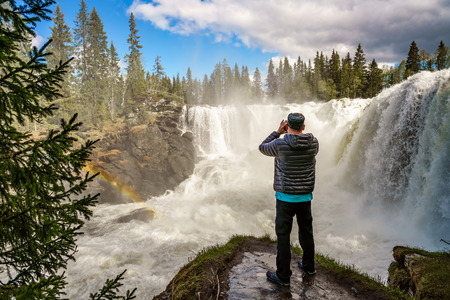Nature photographer tourist with camera shoots while standing Ristafallet waterfall in the western...