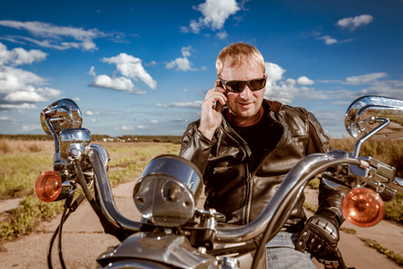Biker talking on a smartphone. Biker man wearing a leather jacket and sunglasses sitting on his motorcycle.の写真素材