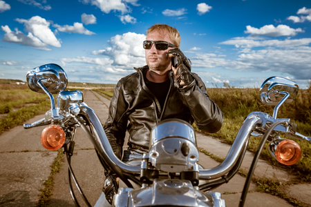 Biker talking on a smartphone. Biker man wearing a leather jacket and sunglasses sitting on his motorcycle.の写真素材