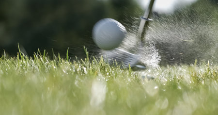Golf club hits a golf ball. Drops of morning dew and grass particles rise into the air after the...