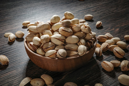 Wooden bowl filled with pistachios on dark wooden table, tasty salted nuts scattered around, healthy...