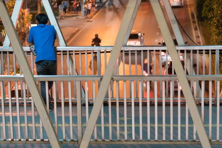 Man standing alone on the bridge. Asian man standing behind and holding a beer can on the footbridge and looking at the car run on the street, in the city night time.の写真素材
