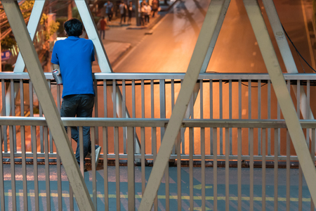 Asian man standing behind and holding a beer can on the footbridge and looking at the empty street not running cars, This is night time in the city of Bangkok.の写真素材
