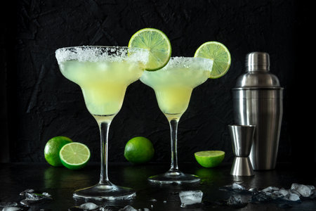 Frozen Margarita cocktails in the classic glasses, side view on a black background, with lime slices and a salt rim, with a shaker and a jiggerの写真素材