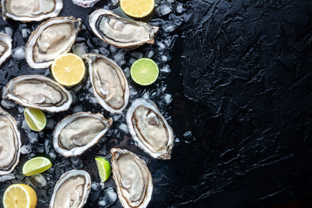 Fresh raw oysters on ice background, overhead flat lay view on a black slate texture, with lemon and lime, with copy spaceの写真素材