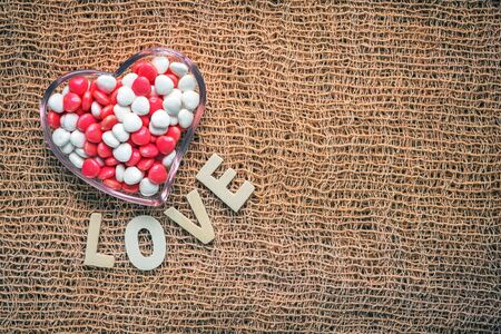 heart shape bowl of color candy and wooden letter love on fabric background.の写真素材