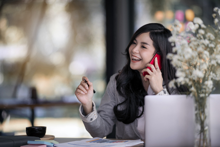 Attractive young asian business woman talking on the mobile phone and smiling while sitting at her working place in office and sharing good business news. の写真素材