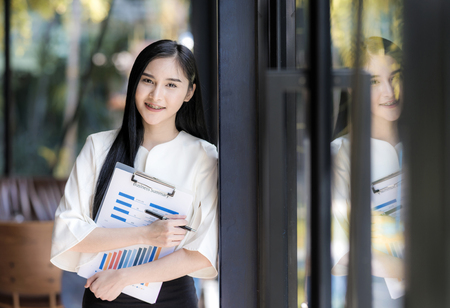 Smiling attractive caucasian businesswoman holding business summary chart standing near glass wall with reflectionの写真素材