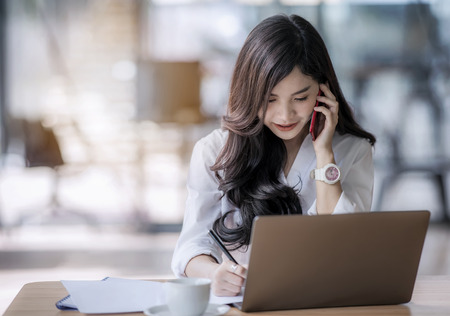 young asian business woman using laptop and writing on notebook, woman officer hard working communicate with customer and recordの写真素材