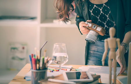 Cropped image of young designer woman working with paperwork and holding digital camera while standing near the desk in her workshopの写真素材