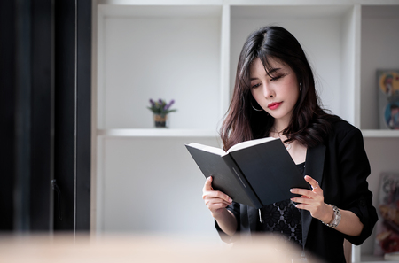 Beautiful woman in black suit sitting and reading a book in white roomの写真素材