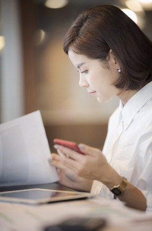 Beautiful businesswoman sitting at desk ,holding smartphone and looking at paper work on her deskの写真素材