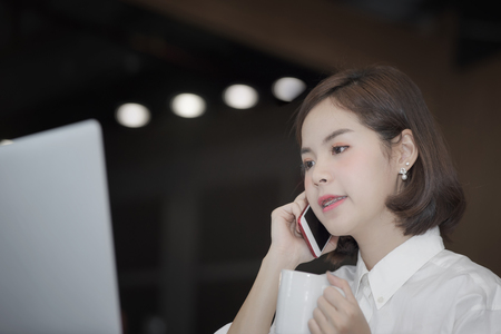 Beautiful businesswoman holding cup of coffee and using smartphone while working at modern office.の写真素材