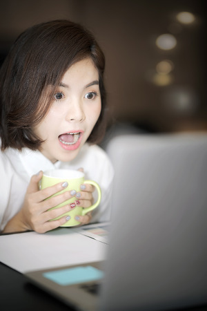 Beautiful businesswoman open wide her mouth and surprised when looking at screen of laptop, sitting and working at her deskの写真素材