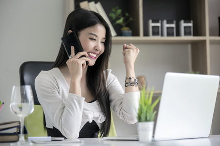 Beautiful asian businesswoman hands up and cheerful while using smartphone and looking at laptop screen in modern office の写真素材