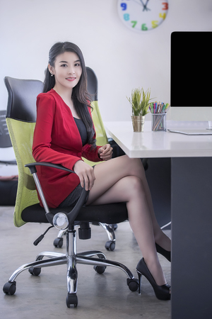 Beautiful businesswoman in red suit smiling and sitting in office, looking at cameraの写真素材