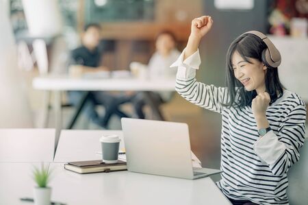 Young businesswoman sitting at an office desk in front of laptop with her hands up, looking forward to business success.の写真素材