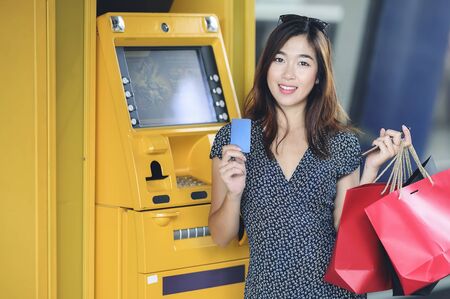 Beautiful asian woman with shopping bags holding credit card standing near yellow ATM machine and looking at camera.の写真素材