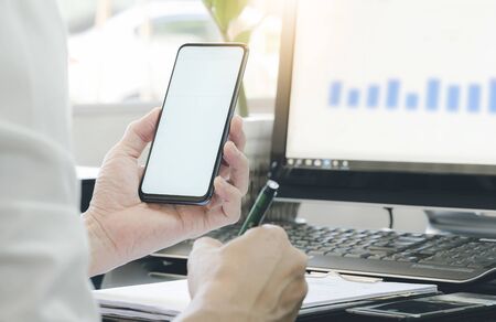 Cropped shot of businessman using smartphone and writing on notepad white working with computer at medern office.の写真素材