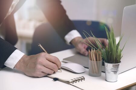 Closeup shot of businessman writing on notebook with pen while standing at office desk in modern office.の写真素材