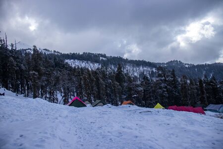 A campsite on a himalayan mountain with tents on the mountain surface and tall pine trees covered with snow in the backgroundの写真素材
