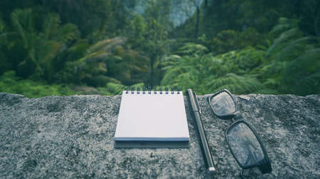 Conceptual  - A notepad, pen and eyeglasses on a concrete table with nature background.の写真素材