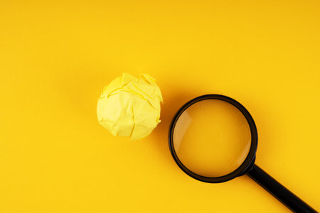 Close-up of yellow crumpled paper ball and a magnifying glass on a yellow backgroundの写真素材