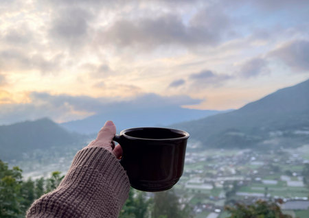 Cozy morning coffee with a scenic mountain sunrise view. Hand holding a warm cup overlooking a peaceful valley. Relaxing travel lifestyle and nature landscape concept.の写真素材