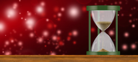 An hourglass with green accents sits on a wooden surface against a red background with blurry white...