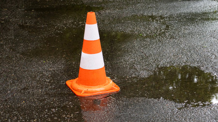 A vibrant orange traffic cone rests on slick pavement, surrounded by small puddles. Rain has fallen...