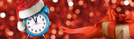 A vibrant blue alarm clock adorned with a Santa hat stands next to a nicely wrapped gift with a red ribbon. The background sparkles with festive red lights, creating a cheerful holiday scene.の写真素材