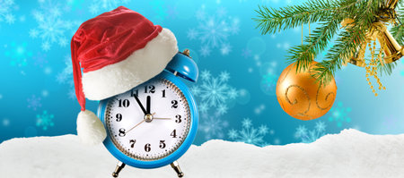 A blue clock wearing a Santa hat sits on a snowy surface surrounded by festive decorations....