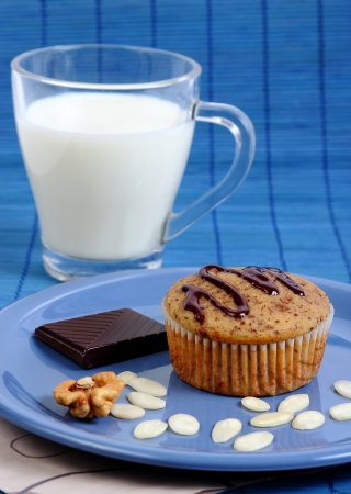 Closeup image of a muffin cake on a blue plate with almond chips, walnut, a piece if dark chocolate and a glass of milkの写真素材