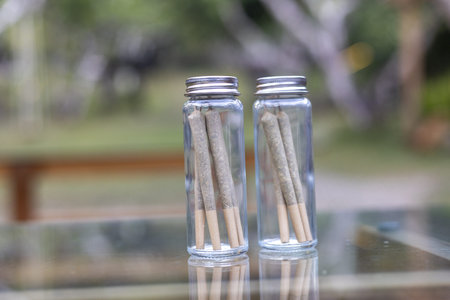 Two glass jars with herbal cannabis joints represent natural recreational marijuana lifestyle. Medical drug for relaxed and calm feeling, with blurred outdoor background