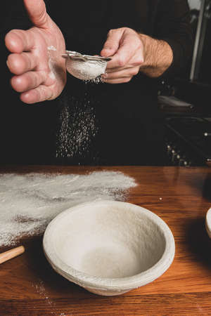 Close up view of baker sifting flour onto wooden bench in preparation for baking bread with banneton...