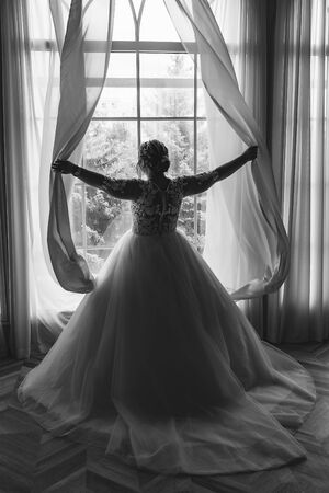 The bride XL in a beautiful white dress opens the curtains with her hands.の写真素材
