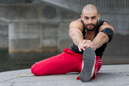Professional fitness athlete trainer. Muscular male sportsman is training himself. Man is doing stretching. Outdoors fitness sport concept. Street flexibility workout in headphonesの写真素材