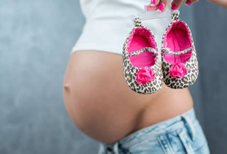 Close up of a cute pregnant belly and little shoes for the baby. Pregnant female motherhood concept....
