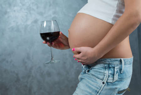 Close up of a cute pregnant belly and glass of wine. Alcohol and pregnancy. Pregnant female unhealthy motherhood concept. Side view, pregnant belly body partの写真素材