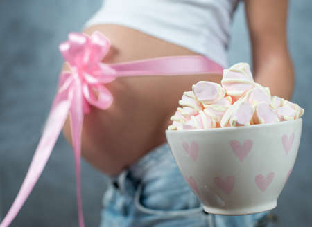 Close up of a cute pregnant belly tummy with pink ribbon and marshmellow. Pregnant female motherhood...