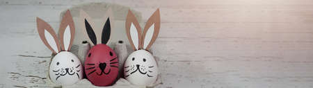 Happy Easter background banner panorama greeting card -Close-up from white and pink cute funny painted eggs and easter bunny in egg carton on rustic white vintage shabby wooden tableの写真素材