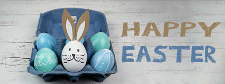 Happy Easter background banner panorama greeting card -Close-up from colorful painted eggs and easter bunny in blue egg carton on rustic white vintage shabby wooden tableの写真素材