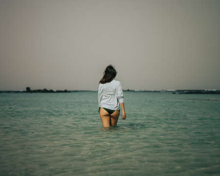 Girl entering the blue, lonely and calm sea of Lanzarote one winter morning while raising her left hand and touching her hair that is blowing in the windの写真素材