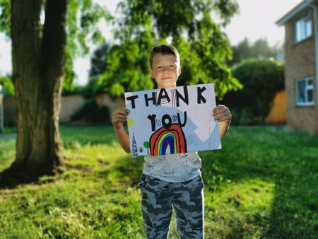 A young boy holds a rainbow drawing and the Thank You message for the medical staff working through the Covid-19 Pandemicの写真素材
