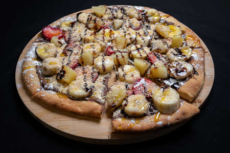 Pizza with chocolate sauce, caramel sauce, strawberries, bananas, pineapple and powdered sugar on a black background.の写真素材