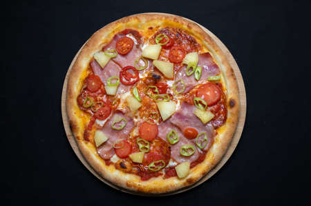 Pizza with tomato sauce, chicken ham, spicy salami, cherry tomatoes, pineapple and hot peppers on a black backgroundの写真素材