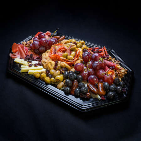 Festive plate with black and white olive mozzarella cheese, grapes, olive salami, sausages and nuts on a black background.の写真素材